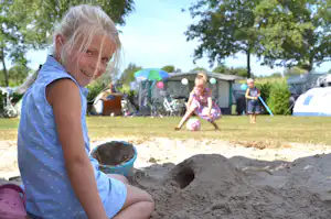 Kind kniet im Sand auf dem Campingplatz, im Hintergrund Zelte, Spielzeug und weitere Kinder im Gartenbereich.