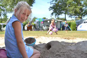 Kleines Kind sitzt im Sand und spielt am Campingplatz, im Hintergrund Zelte und weitere Kinder im Spielbereich.