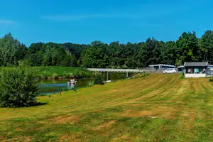Grünes Ufer am See mit Rasenfläche, Steg und Wohnwagen am Campingplatz, im Hintergrund Bäume und Gebäude.