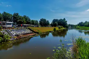 Campingplatz am Fluss: Wohnwagen mit Terrasse, Holzsteg und Liegestühlen am Ufer, dahinter Wiesen und Bäume.