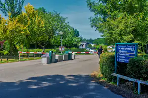 Eingang zum Campingplatz mit Weg, Absperrung und grünen Mülltonnen, im Hintergrund Gebäude und Schilder „’t Wisentbos“.