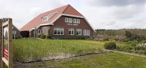 Campinghaus mit rotem Ziegeldach, großen Fenstern und gepflegtem Rasen, im Hintergrund Bäume und ein Weg.
