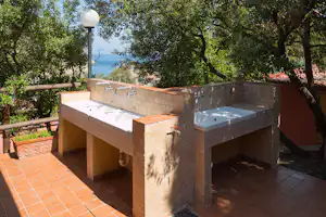 Außenwaschbecken mit Wasserhähnen und Handtuchablage auf einer Terrasse, umgeben von Bäumen und Blick auf das Meer.
