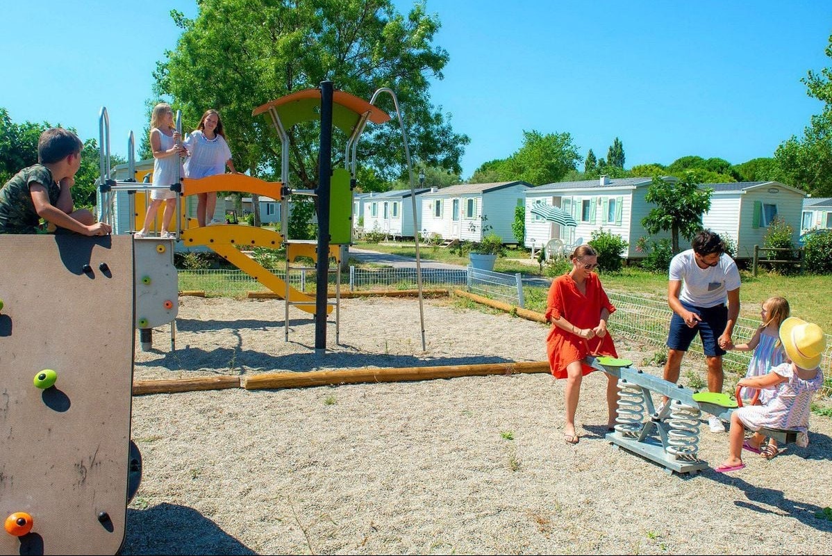 Spielplatz am Campingplatz mit Kletterwand, Sandfläche und Kindern vor Wohnwagen und weißen Sanitärgebäuden.