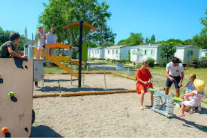 Spielplatz am Campingplatz mit Kletterwand, Sandfläche und Kindern vor Wohnwagen und weißen Sanitärgebäuden.