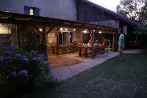Terrasse am Campingplatz mit Holzdach, mehreren Tischen und Stühlen, beleuchteten Lampen und Hauswand im Hintergrund.