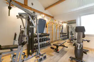 Fitnessraum im Campingbereich mit Kraftstation, Hanteln und Trainingsgeräten, Holzdecke und großen Spiegeln an der Wand.