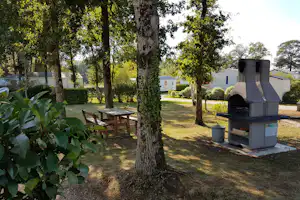Campingplatz-Garten mit Grill, Holztisch und Stühlen zwischen Bäumen, im Hintergrund Wohnwagen und Wege.