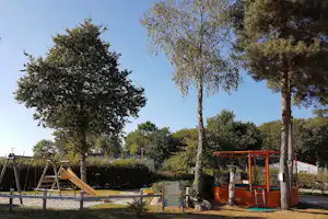 Spielplatz im Campingpark mit Klettergerüst, Schaukel und Bäumen im Hintergrund, Rasenfläche und Zaun sichtbar.