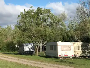 Wohnwagen auf einem Campingplatzstellplatz neben Bäumen, mit Hecke und Schotterweg im Vordergrund.