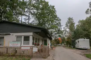 Campinghaus mit Holzlattenzaun, Schild „Springhorstsee GRILLPLATZ“ und Sitzbank vor dem Gebäude, im Hintergrund Bäume und Wohnwagen.