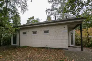 Campinggebäude mit Eingangstür unter Vordach, Ziegelwand und gepflastertem Weg, umgeben von Bäumen und Laub.