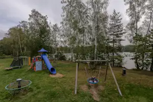 Spielplatz am Campingplatz: Rutsche, Kletterelement und Schaukel auf Rasen, im Hintergrund Bäume und ein See.