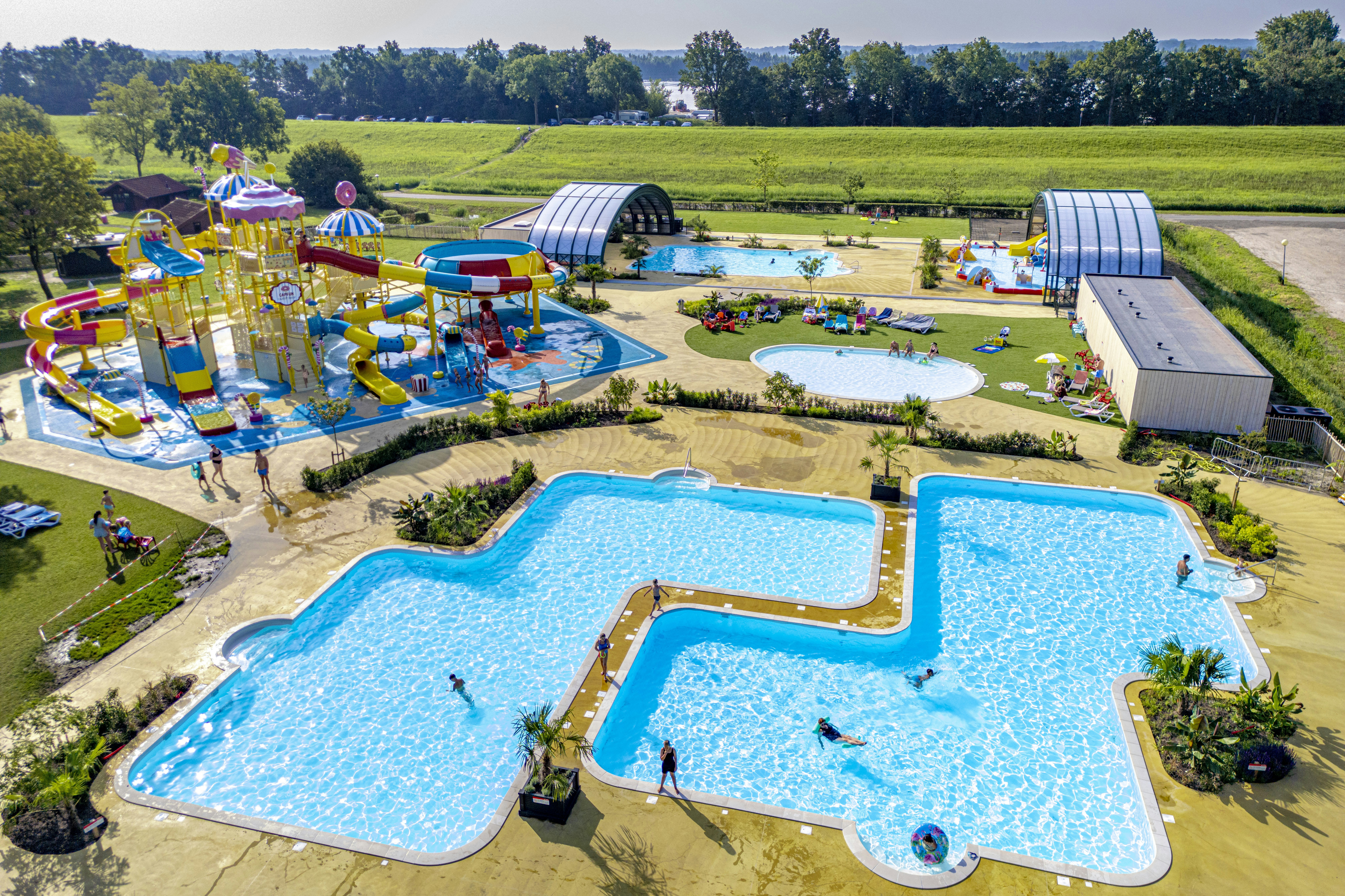 Luftaufnahme eines Campingplatzes mit mehreren Pools, Liegen, Spielbereich und grünen Wiesen, im Hintergrund Bäume und Gebäude.