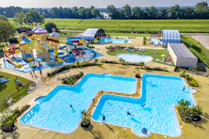 Luftaufnahme eines Campingplatzes mit mehreren Pools, Liegen, Spielbereich und grünen Wiesen, im Hintergrund Bäume und Gebäude.