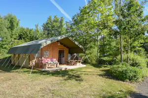 Zelt mit Holzterrasse und Sitzgelegenheiten im Grünen, umgeben von Bäumen und Rasenfläche.