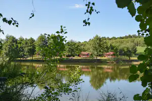 Ruhiger Camping-See mit Spiegelung, Bäumen und zwei roten Ferienhäusern am Ufer, im Vordergrund grünes Blattwerk.