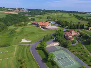 Luftaufnahme einer Campinganlage mit Gebäuden, Innenhof, Tennisplatz und Zufahrtswegen inmitten grüner Hügel.