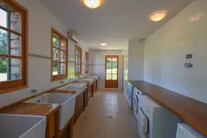 Waschraum im Campingbereich mit mehreren Waschbecken, Holzablagen, Fenstern und weißen Waschmaschinen an der Wand.