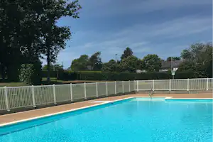 Blaues Schwimmbecken auf dem Campingplatz, umgeben von weißem Zaun, Bäumen und Häusern im Hintergrund.