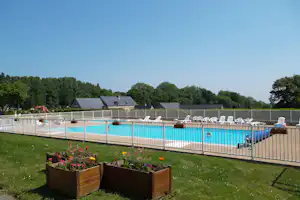 Schwimmbecken mit blauem Wasser am Campingplatz, umzäunt mit Holzgeländer, im Vordergrund Blumenbeete und Rasen.