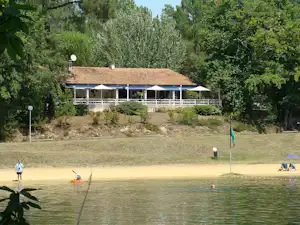 Terrasse eines Gebäudes am See mit Sonnenschirmen, dahinter Bäume und ein gepflegter Uferbereich mit Badenden.