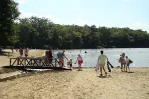 Sandstrand am Campingplatz, Menschen laufen am Ufer entlang, im Hintergrund Bäume und das ruhige Wasser.