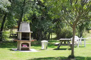 Grillplatz auf dem Campinggelände: gemauerter Grill mit Holz, daneben Bank und weiße Gartenstühle im grünen Garten.