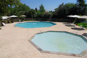 Zwei Außen-Schwimmbecken auf einem gepflasterten Bereich, mit Liegen und Sonnenschirmen, umgeben von Bäumen und Zaun.