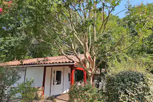 Kleines Ferienhaus mit rotem Ziegeldach, weißer Fassade und Eingangstür, umgeben von Bäumen und Hecken im Garten.