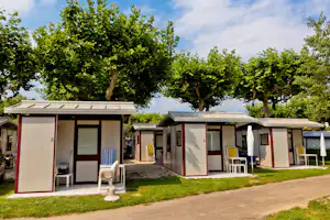 Camping-Bungalows mit Terrasse, Sitzgelegenheiten und Markise, zwischen Bäumen auf einem gepflegten Rasen.