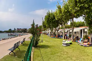 Campingplatz am See: Rasenfläche mit Zelten, Liegestühlen und aufblasbaren Booten, daneben Uferpromenade mit Bäumen.