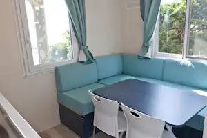 Wohnbereich mit Eckbank und Tisch, weiße Stühle und große Fenster mit grünen Vorhängen, innen auf Holzfußboden.