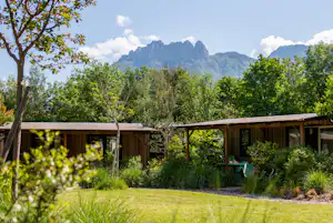 Moderner Holzbungalow mit Terrasse, Sitzbank und Pflanzen im Garten, im Hintergrund Bäume und Berge.