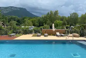 Camping-Pool mit mehreren Liegen am Beckenrand, Zaun und Bäumen im Hintergrund, Berge und bewölkter Himmel.