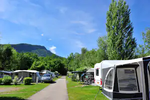 Reihe von Wohnwagen und Zelten auf einem Campingplatz, mit Rasenwegen, Bäumen und Bergen im Hintergrund.