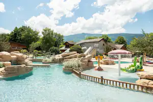 Schwimmbecken mit Holzgeländer, Wasserspiel und bunten Figuren; im Hintergrund Poolbereich, Gebäude und Bäume vor Bergen.