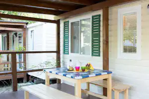 Terrasse am Wohnwagen mit Holztisch, Sitzbank und grünen Fensterläden, überdacht von einer Holzpergola.