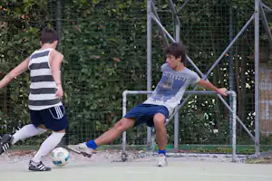 Zwei Männer spielen Fußball auf einem Campingplatz, neben einem Metalltor und einem Kunstrasenfeld mit Zaun im Hintergrund.