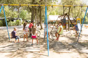 Spielplatz auf dem Campingplatz mit blauen Schaukeln, Sandboden und Spielgeräten zwischen Bäumen und Wegen.