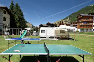 Tischtennisplatte auf dem Campingplatz, daneben Wohnwagen und Zelt, im Hintergrund Häuser und Berge mit Nadelbäumen.