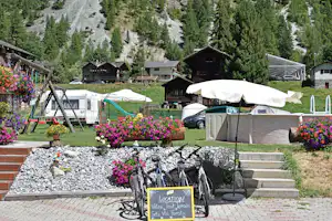 Campingplatz mit Wohnwagen, weißen Markisen und Blumenbeeten vor Stufen, im Hintergrund Berge und Holzchalets.