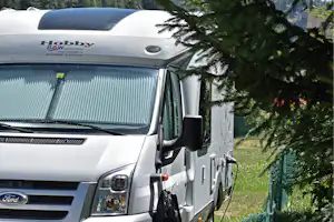 Weißes Wohnmobil am Campingplatz, neben Zaun und Bäumen, mit Blick auf Berge und einen schneebedeckten Gipfel.