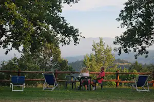 Sitzgruppe auf einer Camping-Terrasse mit Stühlen und Liegen, umgeben von Bäumen, Blick auf Hügel in der Ferne.
