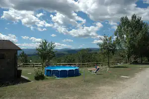 Auf dem Campingplatz steht ein blaues Aufstellpool im Garten, mit Liegestühlen, Bäumen und Blick auf die Hügel.