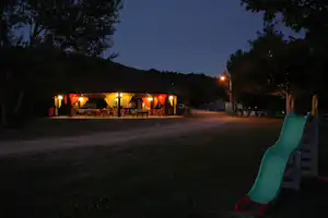 Zeltunterkunft am Campingplatz am Abend mit beleuchteter Terrasse, Tischen und Stühlen, im Vordergrund eine Rutsche.