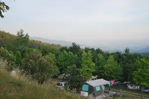 Campingzelt auf einem Stellplatz im Grünen, mit Stühlen und Auto am Rand, Blick über das Tal und bewaldete Hügel.