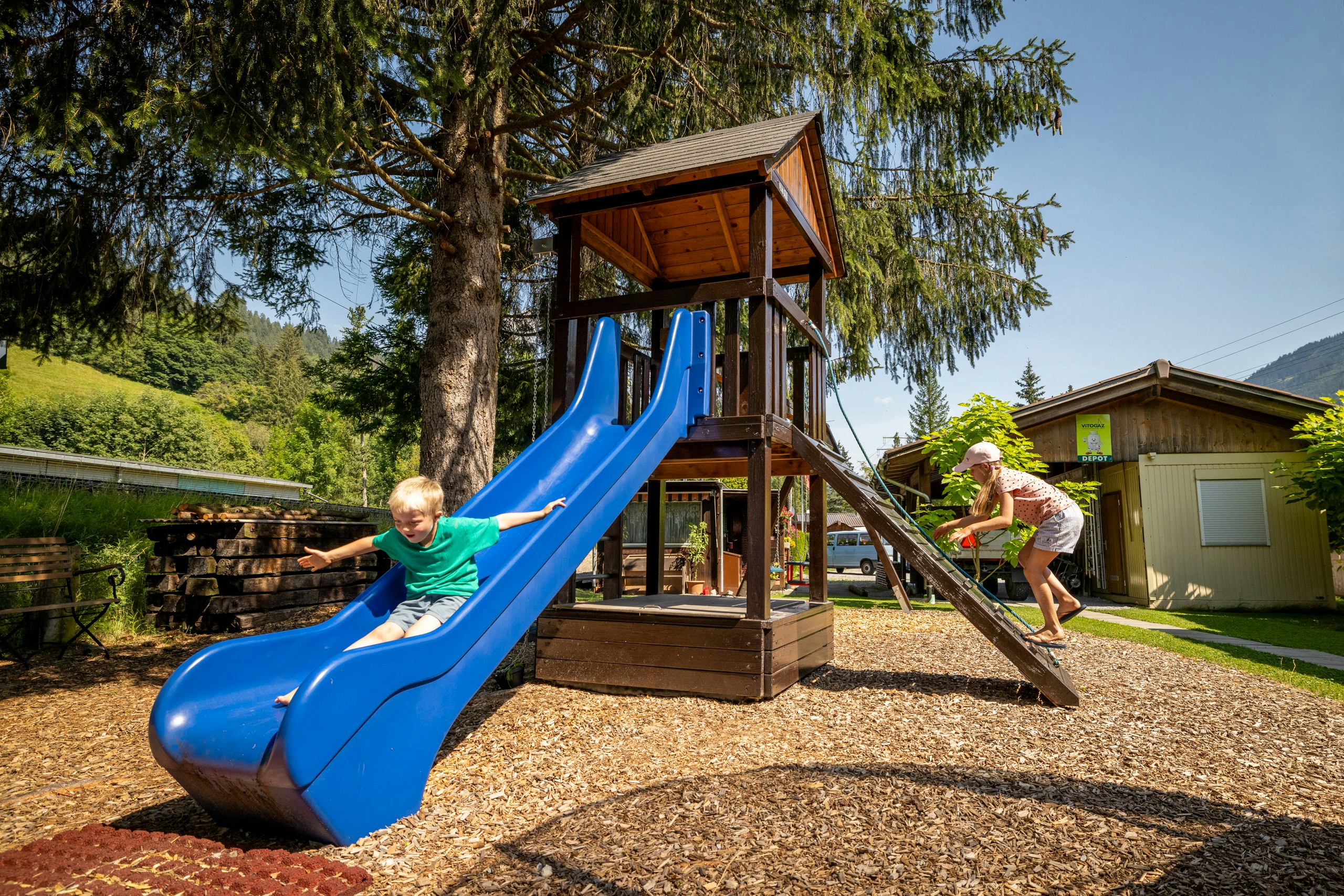 Spielplatz am Campingplatz: Holzspielturm mit blauer Rutsche, zwei Kinder auf dem Steg, Kiesfläche und Bäume im Hintergrund.