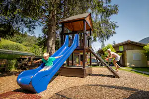 Spielplatz am Campingplatz: Holzspielturm mit blauer Rutsche, zwei Kinder auf dem Steg, Kiesfläche und Bäume im Hintergrund.