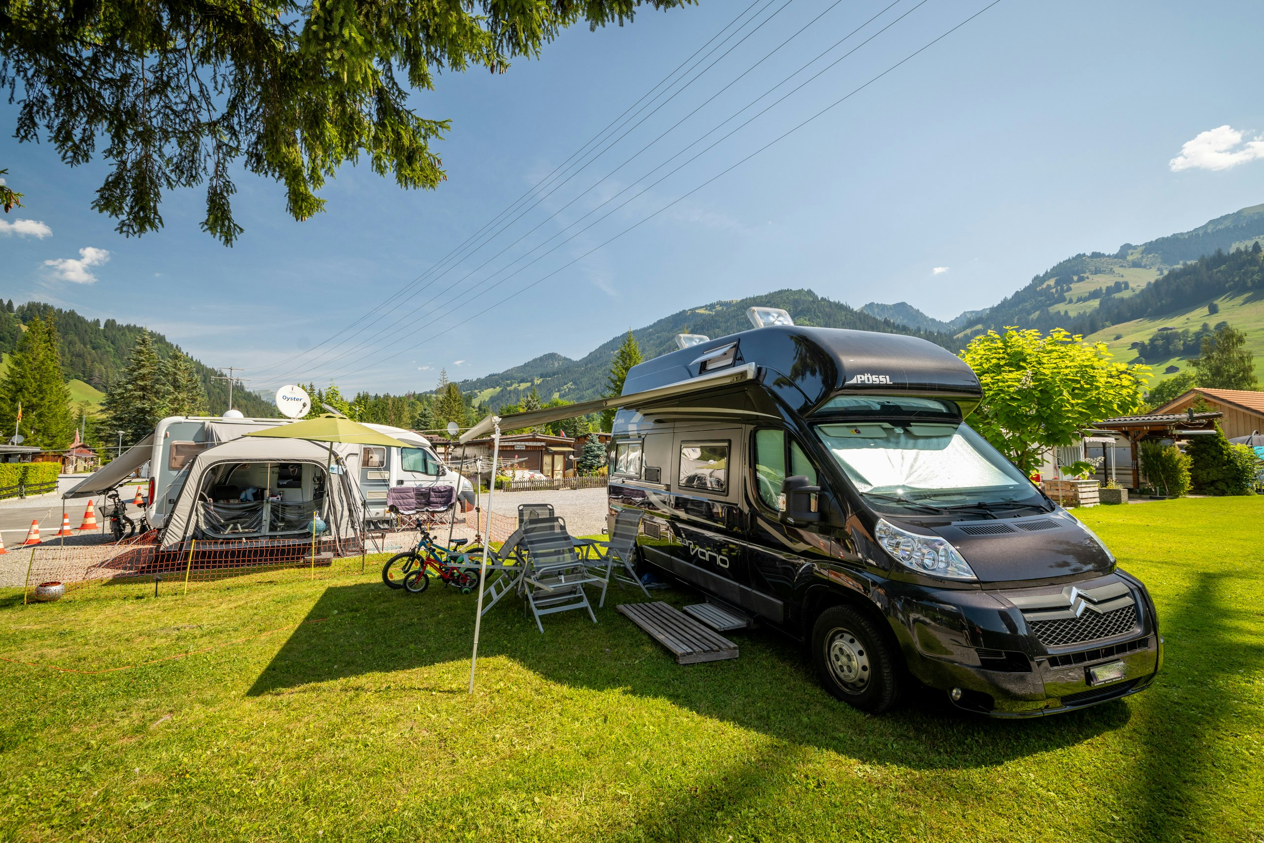 Wohnmobil und Wohnwagen auf einem Campingplatz mit Rasen, Sonnenschirm, Stühlen und Fahrrädern; im Hintergrund Berge und Bäume.